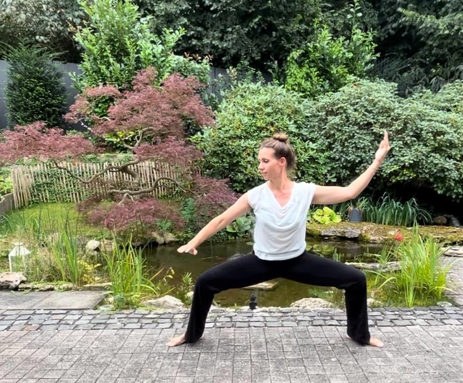 Physiotherapie Claudine König - Heilpraktikerin Physiotherapie - Eine Frau übt eine Yoga-Pose oder eine Tai-Chi-Bewegung in einem üppigen, grünen Garten im Freien.